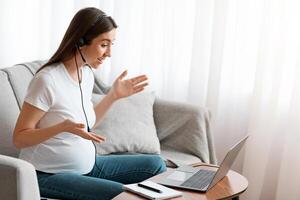 Remote Work. Happy Young Pregnant Female Tutor Making Online Lesson With Laptop, Wearing Headset, Talking And Gesturing At Computer Web Camera, Sitting On Couch In Living Room, Side View photo