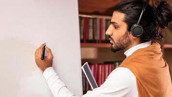 Indian Teacher Man Wearing Headphones Writing English Language Rules On Blackboard Having Virtual Lecture With Distant Students Standing Indoors. Web Tutoring, E-Teaching Concept photo