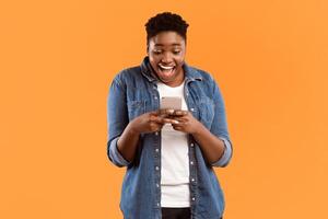 Excited African American Lady Using Cellphone Texting In Social Media Or Playing Mobile Game Standing Over Yellow Studio Background. Great Mobile Application For Your Smartphone Concept photo