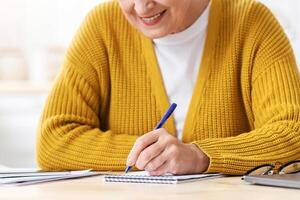 Cropped of old woman sitting at table with papers, laptop and mobile phone in kitchen, taking notes, making shopping list or counting month spendings. unrecognizable woman writing in notepad photo
