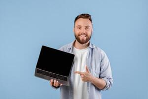 Positive young man pointing at empty laptop screen with space for your website design over blue studio background, mockup. Millennial guy showing PC with space for your advertisement photo