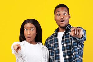 Wow, what is this. This is unimaginable and amazing. Shocked teen african american students man and lady in casual with wide open eyes pointing fingers at camera, isolated on yellow background photo