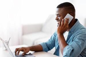 Work From Home. Smiling Black Freelancer Guy Using Laptop And Cellphone, Young African American Man Talking On Mobile Phone And Working Online On Laptop Computer At Home Office, Copy Space photo