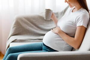 Smiling young pregnant lady relaxing on couch with cup of herbal tea, positive expecting woman enjoying hot drink while resting on sofa in living room, spending time at home, side view with copy space photo