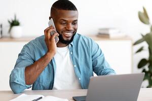 Remote Business. Smiling Black Male Freelancer Talking On Cellphone And Working On Laptop At Home Office, African Man Using Computer And Having Pleasant Phone Conversation, Enjoying Distance Job photo
