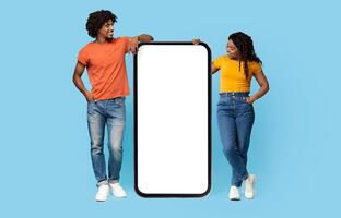 Curious millennial african american couple in casual outfits looking at huge smartphone with white empty screen between them and smiling, mockup, blue studio background, full length shot photo