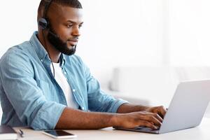Black millennial man in headset working remotely with laptop at home office, young african american freelancer guy using computer, typing on keyboard while sitting at desk near window, copy space photo