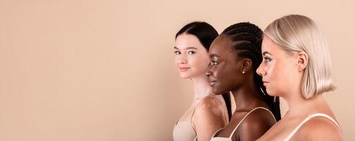Side View Portrait Of Three Beautiful Interracial Women. Group Of Young Attractive Body Positive Females With Different Skin Types Wearing Lingerie Standing Over Beige Studio Background, Free Space photo