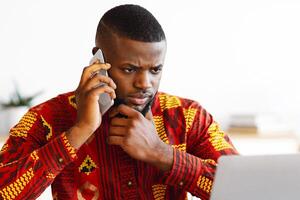 Portrait Of Busy Black Businessman In Tribal Shirt Talking On Cellphone And Working On Laptop, African Entrepreneur In Traditional Clothes Looking At Computer Screen And Having Phone Conversation photo