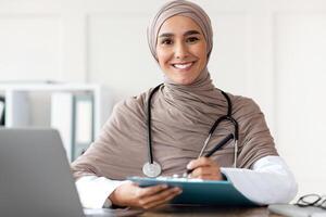 Beautiful muslim woman doctor writing in medical chart and smiling at camera, wearing hijab, sitting at workdesk with modern laptop in her cabinet in clinic or hospital, taking anamnesis, copy space photo