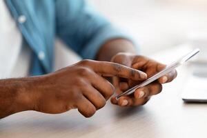 Cropped Image Of Unrecognizable Black Man Using Smartphone While Sitting At Desk In Office, African American Guy Holding Cellphone Browsing New Mobile App Or Shopping Online, Closeup Shot photo