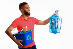 African American Guy Posing With Box Full Of Recycling Plastic And Blue Garbage Bag Standing Over White Background In Studio. Sort And Recycle Your Waste Concept, Side View photo