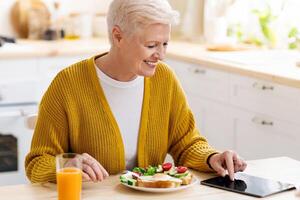 Smiling old lady modern grandmother using digital tablet while having lunch, eating sandwich and drinking juice, sitting alone at kitchen, empty space. Senior woman using newest app on pad photo