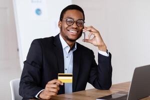 Easy payments. Happy african american businessman holding credit card after online purchase and talking on mobile phone with customer service while working with laptop in his office, free space photo