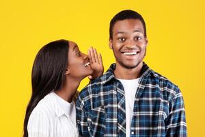 Big secret, sharing news. Mysterious millennial african american student lady in casual whispers in ear of smiling guy something interesting, isolated on yellow background, empty space, studio shot photo