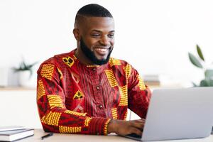 Successful Black Businessman In African Traditional Clothes Sitting At Workplace In Office, Happy Male Entrepreneur In Ethnic Shirt Working With Laptop Computer At Desk, Typing On Keyboard And Smiling photo