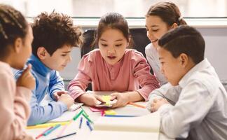 Diverse Group of Cute Small Children Using Smartphone, Sitting Together At Desk In Classroom Having Fun. Multicultural Kids Browsing Internet and Playing Online Games on Mobile Phone photo