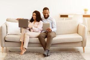Weekend Leisure. Portrait Of Happy Millennial Black Couple Resting On Sofa. Man Using Digital Tablet, Woman Working On Laptop Computer. Family Browsing Internet Together Sitting On Couch photo