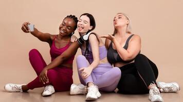 Cheerful Diverse Multiethnic Females In Sportswear Taking Selfie On Smartphone After Training, Happy Body Positive Females Having Fun Together, Sitting On Floor On Beige Studio Background, Free Space photo