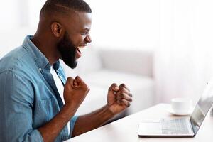 Overjoyed african american freelancer man looking at laptop and cheering success. Excited black guy sitting at desk with computer at home, raising fists and exclaiming with joy, side view, copy space photo