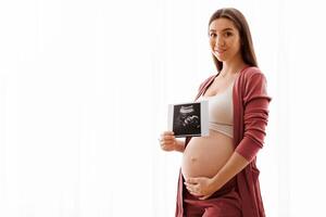 Cute Pregnant Lady Posing With Baby Sonography Photo Near Window At Home, Beautiful Expectant Lady Tenderly Touching Her Belly And Smiling At Camera, Enjoying Healthy Pregnancy And Future Maternity