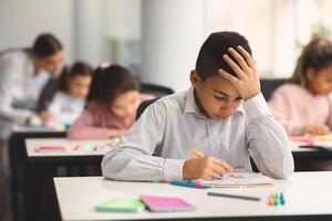 Education And Learning Concept. Portrait of focused small boy sitting at desk in classroom at school, writing in notebook, having test or exam and thinking, touching and grabbing head photo