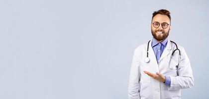 Cheerful male doctor in uniform pointing at empty space over grey studio background, panorama. General practitioner looking at camera and offering medical services, banner design photo