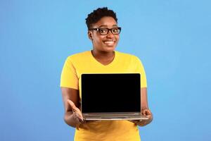 Smiling Oversized Black Woman Holding Laptop With Blank Screen Posing Over Blue Studio Background. Lady Showing Computer Monitor Recommending Website. Mockup photo