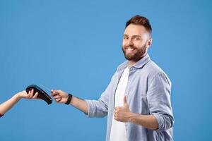 Happy millennial guy using credit card and payment terminal, showing thumb up gesture on blue studio background. Positive young man recommending contactless shopping during covid-19 outbreak photo