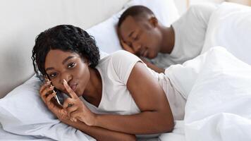 Black woman talking with lover on mobile phone in bed while husband sleeping, cheating in relationships concept. African american woman showing shh gesture at camera while having conversation on phone photo