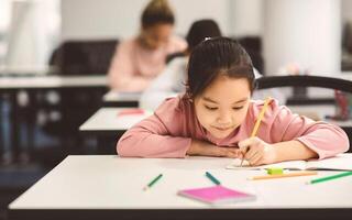 Tuition Concept. Portrait of concentrated small asian girl sitting at table in classroom, writing or drawing in notebook, doing homework or taking notes during lesson, selective focus, free copy space photo