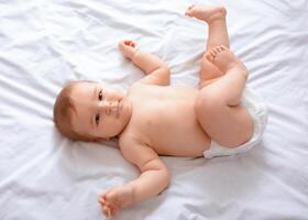 Top view of beautiful caucasian baby in diaper laying on bed, smiling at camera, copy space. Adorable toddler in nappy having fun alone, lying on white, laughing. Toddler, baby, infant photo