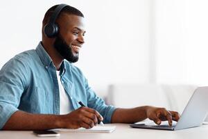 Online Education. Happy Black Guy In Headphones Study In Internet, Watching Webinar On Laptop Computer And Taking Notes, African Man Enjoying Distance Learning, Sitting At Desk At Home, Side View photo