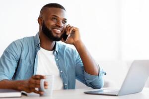 Successful black freelancer guy having coffee break at workplace in home office, smiling african american man talking on cellphone while working on laptop at desk, enjoying conversation, copy space photo