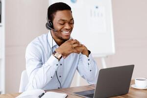 Happy black man having online training, using headset and laptop, looking at computer screen and smiling, office interior, copy space. Cheerful african guy having online call with clients photo