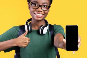 Joyful Black Female Student Showing Smartphone With Empty Screen To Camera And Gesturing Thumbs Up Recommending Application For Cellphone Standing On Yellow Studio Background. I Like This App. Mockup photo