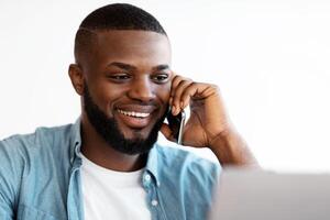 Closeup Portrait Of Smiling Black Man Talking On Cellphone And Using Laptop In Office. Handsome African American Entrepreneur Enjoying Phone Conversation While Working On Computer, Selective Focus photo