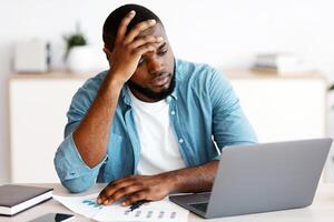 Lack Of Inspiration. Stressed Black Freelancer Guy Sitting At Desk With Laptop And Touching Head In Despair, Having Problems With Project While Working Remotely At Home, Looking At Computer Screen photo