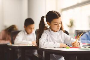Tuition Concept. Portrait of concentrated small girl sitting at table in classroom, writing or drawing in notebook, doing homework or taking notes during lesson, selective focus, free copy space photo