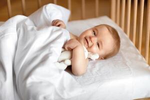 Adorable smiling blonde baby laying in wooden crib under blanket. Happy little kid infant woke up after sleeping, hugging toy and looking at camera, lying in baby bed, close up photo