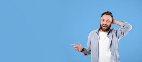 Happy young guy pointing at empty space, cannot believe his luck over huge sale on blue studio background, banner design with copy space. Handsome bearded man promoting your product photo