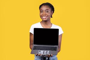 Cheerful Black Lady Showing Laptop Blank Screen Holding Computer Standing Posing On Yellow Studio Background, Smiling To Camera. African Woman Recommending Website. Mockup photo