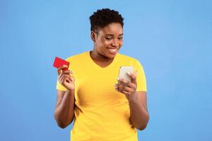 African Lady With Smartphone And Credit Card Using Mobile Banking Application Shopping Online Standing Over Blue Studio Background. Black Woman Using Phone App Concept photo