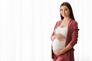 Portrait Of Beautiful Pregnant Woman Standing Near Window At Home, Embracing Belly. Young Expectant Mother Tenderly Touching Her Tummy And Smiling At Camera, Enjoying Pregnancy Time, Copy Space photo