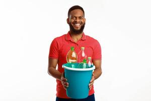 Black Young Male Holding Bucket With Wasted Plastic Bottles Standing Posing Over White Background In Studio, Smiling To Camera. Waste Sorting And Recycling photo