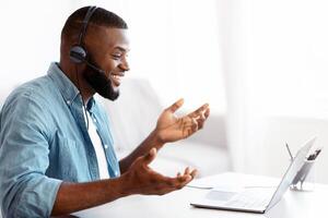 Web Conference. Friendly black guy in headset making call on laptop, african american man sitting at desk in home office, talking at computer camera, enjoying online communication, side view photo