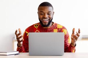 Online Communication. Positive African Guy In Traditional Shirt Having Call On Laptop, Friendly Black Man Sitting At Desk With Computer, Talking And Gesturing At Camera, Copy Space photo