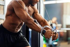 Young black man athlete training triceps on block exerciser in gym, side view, copy space. Shirtless african american bodybuilder having workoout at modern gym, using crossover machine photo