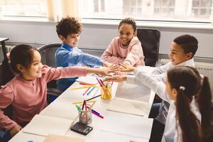 Childhood Support Concept. Group of multicultural emotional junior schoolkids sitting in circle at desk in modern classroom and putting hands together, having fun, studying or playing game photo