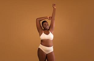 Portrait Of Beautiful Curvy African American Lady Posing Isolated On Brown Studio Background, Black Lady In Beige Underwear Enjoying Herself, Standing With Raised Arms And Closed Eyes, Free Copy Space photo
